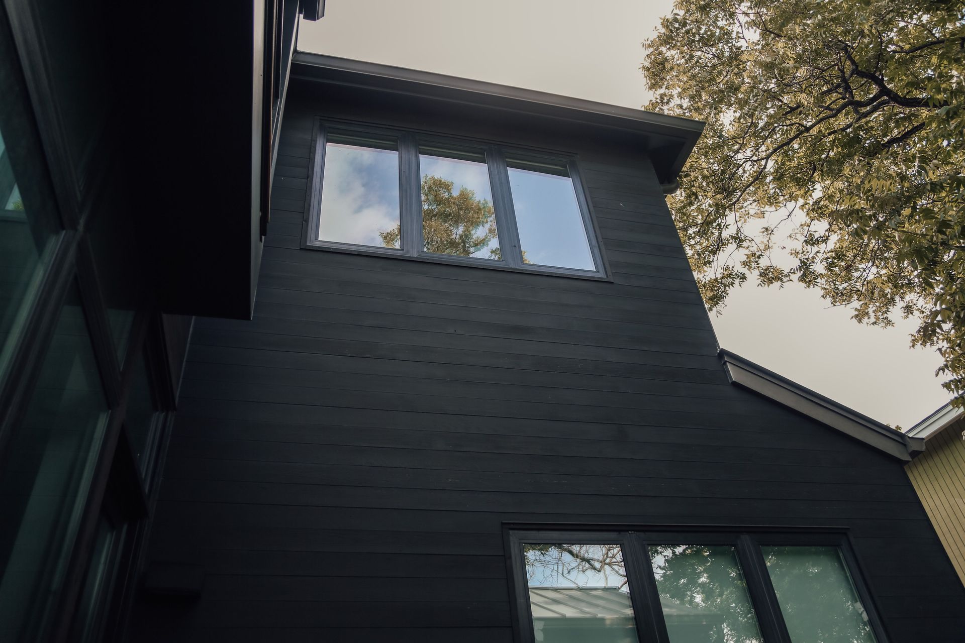From below of gray sky and green foliage over tall black house with small windows and black wooden walls in daytime